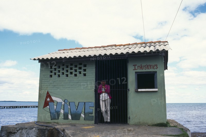 CUBA, West Coast, Vinales,Woman standing in doorway of drinks store building with Vive painted on exterior. The ocean behind.