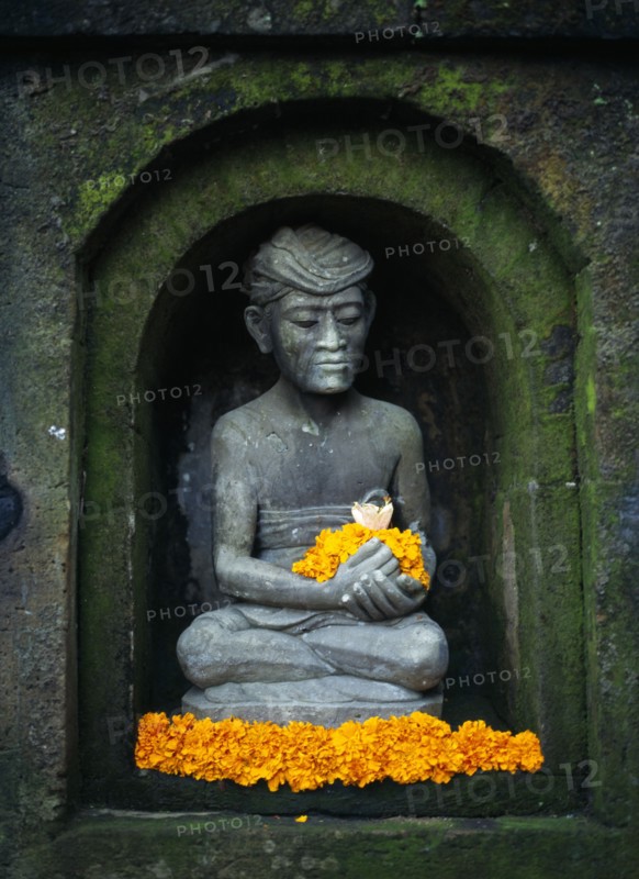 Indonesia, Bali, Ubud, Stone statue of male figure in niche of wall sitting in lotus position holding and surrounded by yellow frangipani flowers.