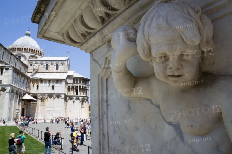 ITALY, Tuscany, Pisa, The Campo dei Miracoli or Field of Miracles with the Duomo Cathedral and tourists under a blue sky with a marble cherub carved into a marble plinth in the foreground.
