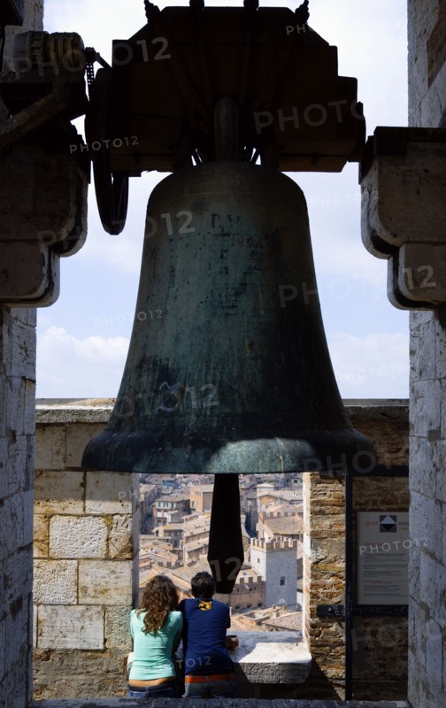 ITALY, Tuscany, Siena, Palazzo Publico Two young tourists at the top of the Torre del Mangia campanile belltower beneath a large bronze bell looking out over the city.