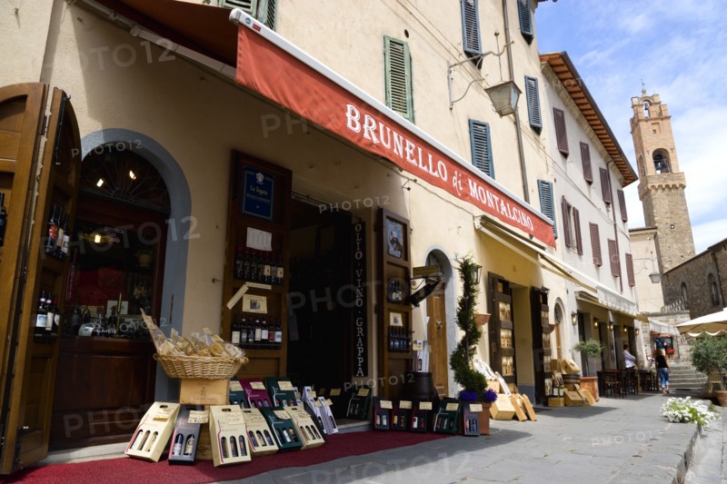 ITALY, Tuscany, Montalcino, Val D'Orcia Brunello di Montalcino Enoteca or wine shop with dispaly of boxed wines on the pavement around its entrance beneath a sunshade. The 14 the Century belltower of the Palazzo Comunale is at the end of the street.