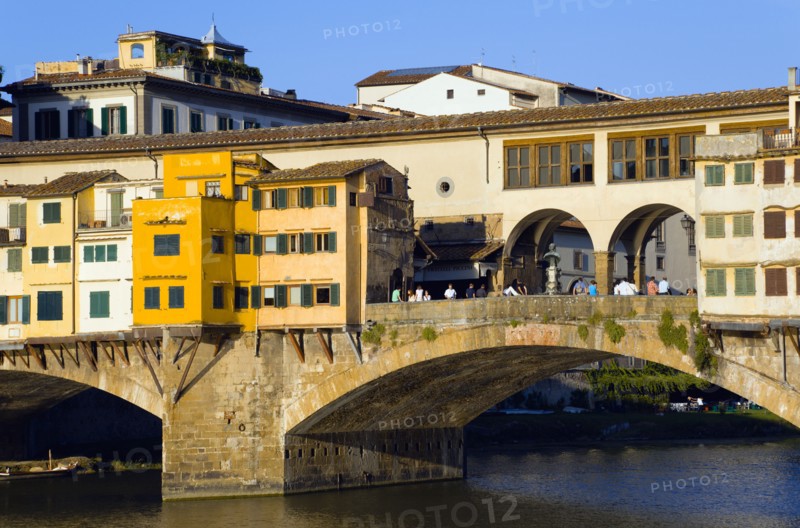 ITALY, Tuscany, Florence, Ponte Vecchio medieval bridge across River Arno with sightseeing tourists beside merchant's shops that line bridge and hang over the water below.