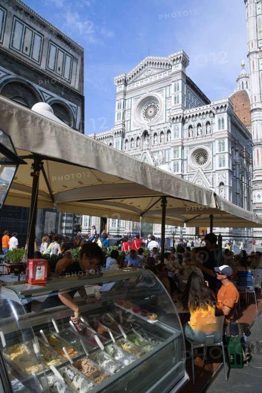 ITALY, Tuscany, Florence, The Neo-Gothic marble west facade of the Cathedral of Santa Maria del Fiore the Duomo with sightseeing tourists people sitting under umbrellas in the shade at a restaurant and a waitress serving ice cream from a counter.