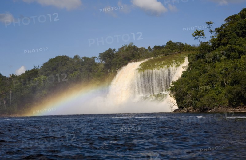 VENEZUELA, Bolivar State, Canaima National Park, Canaima Village, waterfalls feeding into Canaima lake while the humidity of its waters are producing a rainbow effect with blue sky and white clouds.