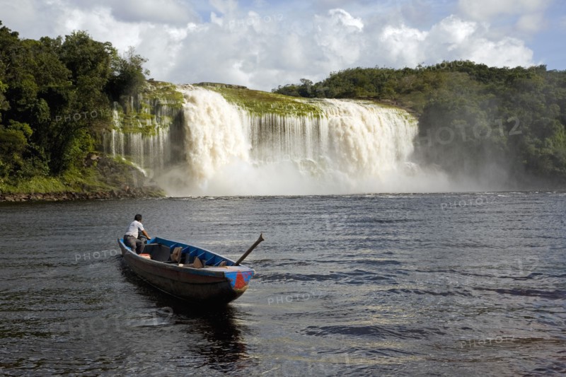 VENEZUELA, Bolivar State, Canaima National Park, Canaima Village, A Pemon tribe man while sailing with his boat at Canaima lake just in front of a big waterfall on a bright day with blue sky and white clouds.