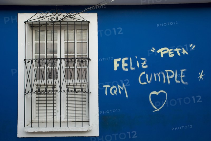VENEZUELA, Bolivar State, Ciudad Bolivar, blue wall with graffiti words in Spanish and the symbol of a heart just next of a closed symmetrical window within a white painted frame, secured with iron bars.