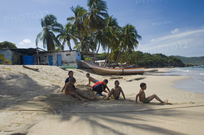 VENEZUELA, Margarita Island, Playa la Galera, Kids playing on the beach, under the shade of a palm tree just in front of the seawater while other trees, a house and a boat are noticeable at the background, shoot on a bright day with blue sky and some white clouds.