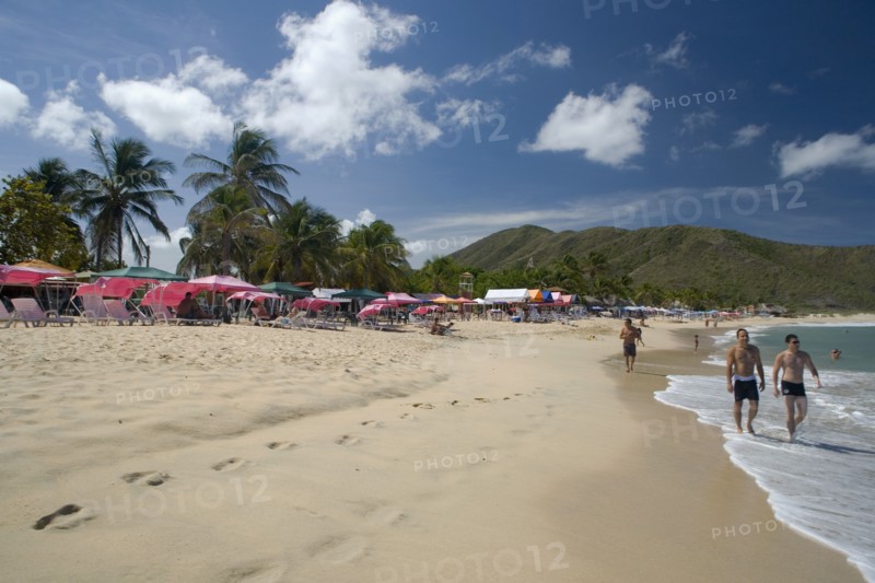 VENEZUELA, Margarita Island, Playa Caribe, View of exotic beach with people walking on the sand while colorful tents, deck chairs, bars, restaurants and palm trees are noticeable, on a bright day with blue sky and white clouds.