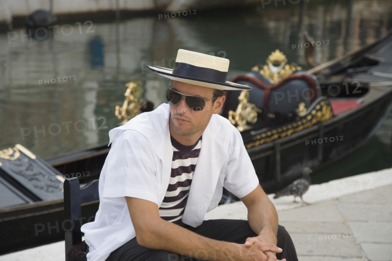Venice, Veneto, Italy. Centro Storico Young gondolier in traditional uniform of striped shirt and straw hat with black sash wearing sunglasses seated beside moored gondola on canal in late summer sunshine. Italy Italia Italian Venice Veneto Venezia Europe European City Sun Shades Sunglasses Canal Water Boater Hat Man People Gondolier Gondolas Gondola Storico Centro Classic Classical Destination Destinations Historical Immature Male Men Guy Older One individual Solo Lone Solitary Southern Europe