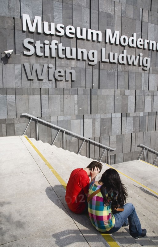 Vienna, Austria. Neubau District. Young student couple sitting on the steps of the Museum of Modern Art Museum Moderner Kunst Stiftung Ludwig Wien or MUMOK. Austria Austrian Republic Vienna Viennese Wien Europe European City Capital Neubau District Quarter Museums Mueseumsquartier Luwig Stiftung Kunst Moderner Museum Facade Exterior Architecture Mumok Couple Color Destination Destinations Gray Immature Osterreich Viena Western Europe