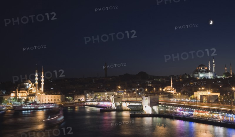 Istanbul, Turkey. Sultanahmet. The Golden Horn. The New Mosque or Yeni Camii at left the Galata Bridge and Suleymaniye Mosque at right illuminated at night with boats on the Bosphorus in motion blur and coloured lights reflected in the water. Turkey Turkish Istanbul Constantinople Stamboul Stambul City Europe European Asia Asian East West Urban Skyline Destination Travel Tourism Sultanahmet Golden Horn New Mosque Yeni Camii Galata Bridge Suleymaniye Dusk Sunset Night Illuminated Skyline Minarets Water Citiscape Building Buildings Urban Architecture Colored Destination Destinations Middle East Nightfall Twilight Evenfall Crespuscle Crespuscule Gloam Gloaming Nite South Eastern Europe Sundown Atmospheric Turkiye Western Asia
