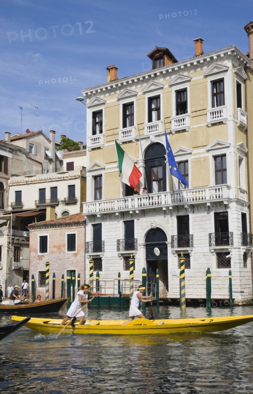 Venice, Veneto, Italy. Female pair taking part in Regata Storica Venice Regatta rowing yellow gondola on the Grand Canal passing waterside buildings with Venice Republic EU and Italian tricolour flags hanging from facades. Italy Italia Italian Venice Veneto Venezia Europe European City Regata Regatta Rowing Gondola Boat Architecture Facade Exterio Flag Flags Water Women Girls Female Blue Destination Destinations European Union Female Woman Girl Lady Southern Europe