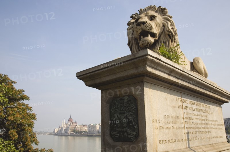 Budapest, Pest County, Hungary. Lion sculpture on the Chain Bridge with Hungarian Parliament Building on far Pest bank in background. Hungary Hungarian Europe European East Eastern Buda Pest Budapest City Transport Architecture Art Lion Statue Carving Sculpture Chain memory Bridge Parliament Building River Danube Destination Destinations Eastern Europe Parliment