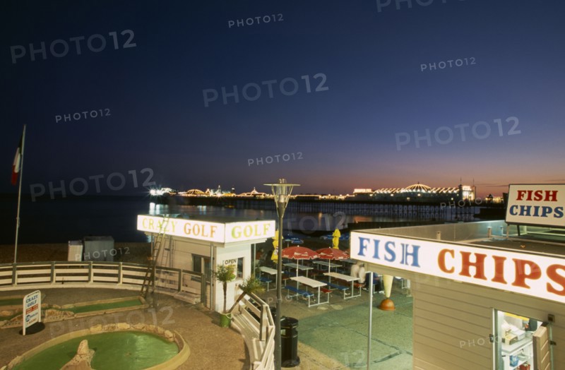 Brighton, East Sussex, England. Crazy Golf and Fish and Chip shop on seafront illuminated at night with Pier in the distance. European Great Britain Nite Northern Europe Store UK United Kingdom British Isles
