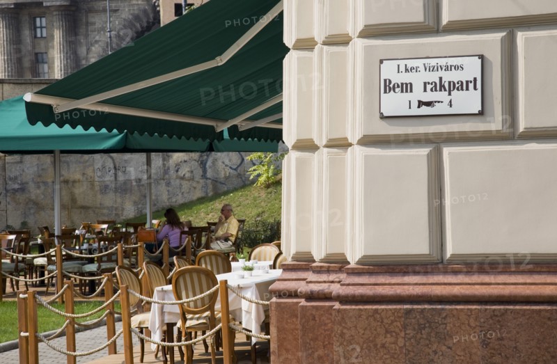 Budapest, Pest County, Hungary. Buda cafe beside the River Danube at approach to Chain Bridge with people seated at outside tables. Hungary Hungarian Europe European East Eastern Buda Pest Budapest City Cafe People Sat Outside Exterior Tables Bar Bistro Destination Destinations Eastern Europe Restaurant