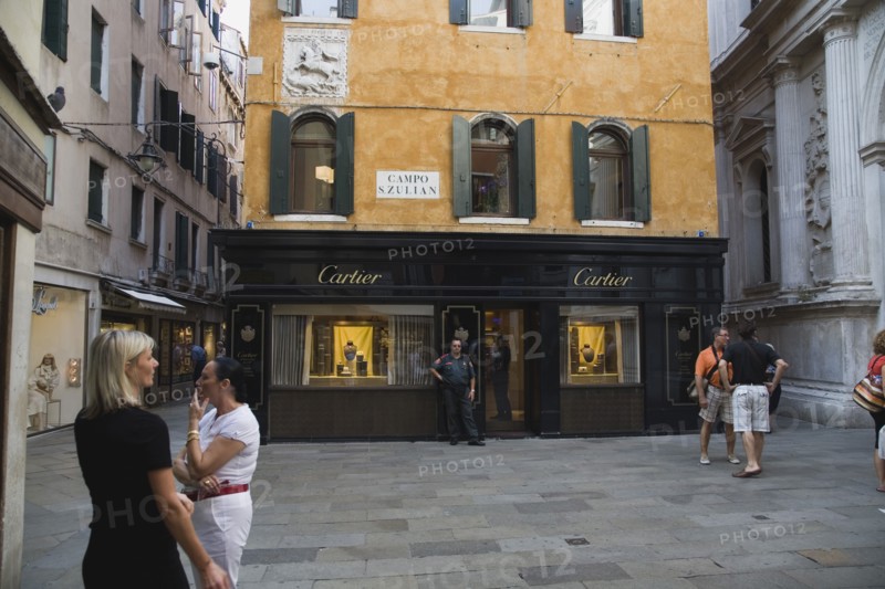 Venice, Veneto, Italy. Centro Storico Cartier jewellery store exterior with security guard and tourists in paved square with shuttered facade and plaque depicting St George and the dragon. Italy Italia Italian Venice Veneto Venezia Europe European City Centro Storico Cartier Jewellery Jewllery Store Shop Campo S.Zulian Square Tourist Tourists People Security Guard Guards Destination Destinations Holidaymakers Jewelry Sightseeing Southeast Asia Southern Europe Tourism