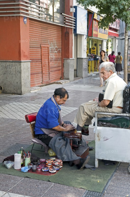 Santiago, Chile. Elderly man at street shoe shine in downtown shopping area piles of shoe polish tins at side. Chilean South America American Hispanic Santiago City Cityscape Urban Street Polish Shoes Shoe Shining Shine Cleaning Clean Working Work Men Man Cleansing Destination Destinations Latin America Latino Male Man Guy Male Men Guy Old Senior Aged Shops Shoppers Mall Retail Buy Buying Market Markets South America Southern Washing