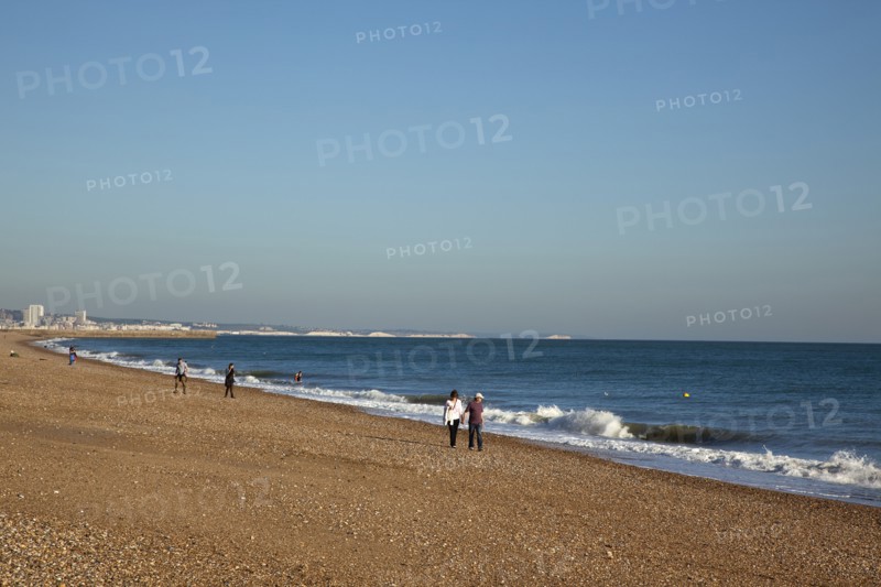 Shoreham-by-Sea, West Sussex, England. People walking along the waters edge on Shoreham beach. England English UK United Kingdom GB Great Britain British Europe European West Sussex County Shoreham Shoreham-by-Sea By Sea Beach People Couple Couples Walk Walking Stroll Strolling Shore Pebble Pebbles Blue Sky