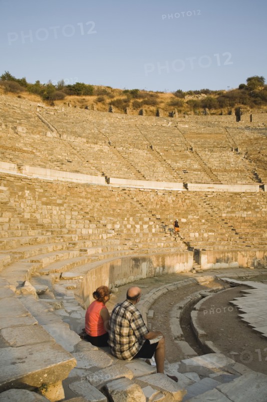Selcuk, Izmir Province, Turkey. Ephesus. Tourist couple sitting in amphitheatre in the antique city of Ephesus on the Aegean sea coast. Turkey Turkish Eurasia Eurasian Europe Asia Turkiye Izmir Province Selcuk Ephesus Ruin Ruins Roman Amphitheatre Aamphitheater Ancient Architecture Masonry Rock Stone Tourists Destination Destinations European History Historic Holidaymakers Middle East Sightseeing South Eastern Europe Tourism Water Western Asia