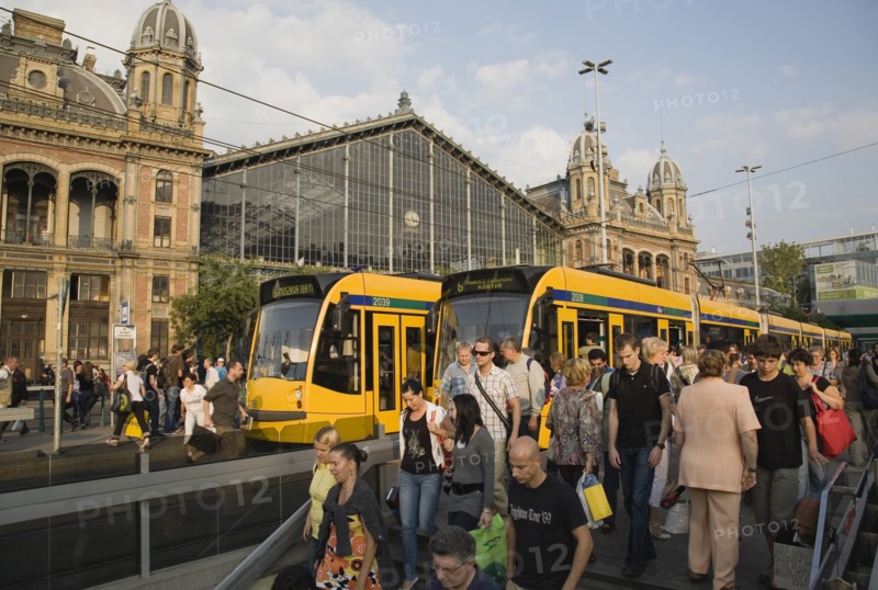 Budapest, Pest County, Hungary. Trams and crowd of passengers in front of rail terminus Budapest Nyugati palyaudvar and walking down steps to metro in foreground. Man appears to be begging on right hand side of steps unacknowledged by passers-by. Hungary Hungarian Europe European East Eastern Buda Pest Budapest City Transport Rail Train Tram Station Terminus Stop Nyugati Palyaudvar People Crowd Crowded Commuters Tourists Travel Passengers Descend Metro Underground Destination Destinations Eastern Europe Holidaymakers Male Men Guy Roundel Tourism