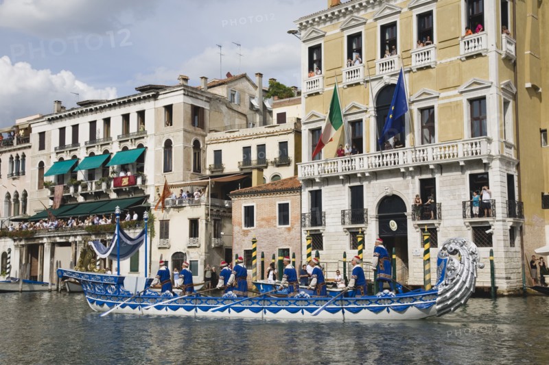 Venice, Veneto, Italy. Participants in the Regata Storico historical Regatta held each September wearing traditional costume and rowing ornately decorated blue and silver gondola approaching the Rialto bridge with onlookers watching from the balconies of canalside buildings behind. Teams represent Sestiere districts of Venice in traditional races. Italy Italia Italian Venice Veneto Venezia Europe European City Regata Regatta Gondola Gondola Gondolas Gondolier Boat Architecture Exterior Water Classic Classical Clouds Cloud Sky Destination Destinations History Historic Holidaymakers Older Southern Europe Tourism Tourist