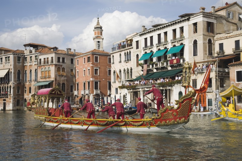 Venice, Veneto, Italy. Participants in the Regata Storico historical Regatta held each September in red and gold decorated gondola wearing traditional costume approaching the Rialto bridge with onlookers gathered on the balconies of canalside buildings behind. Teams represent Sestiere districts of Venice in traditional races. Italy Italia Italian Venice Veneto Venezia Europe European City Regata Regatta Gondola Gondola Gondolas Gondolier Boat Architecture Exterior Water Blue Classic Classical Clouds Cloud Sky Destination Destinations History Historic Holidaymakers Older Southern Europe Tourism Tourist