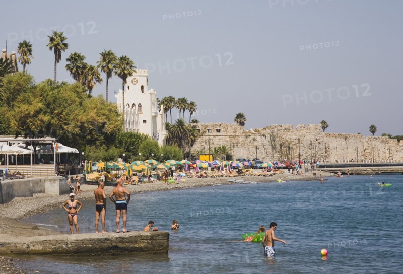 Kos, Dodecanese Islands, Greece. Kos Town beach with castle walls and harbour entrance behind and Greek holidaymakers bathing on shingle beach in foreground. Greece Greek Europe European Vacation Holiday Holidays Travel Destination Tourism Ellas Hellenic Dodecanese Kos Beach Castle Walls Swimmimers Swimming Sunbtherrs Sunbathing Travel Castillo Castello Destination Destinations Ellada Sand Sandy Beaches Tourism Seaside Shore Tourist Tourists Vacation Southern Europe Sunbather