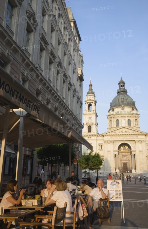 Budapest, Pest County, Hungary. Cafe with Summer tourists seated at outside tables with Saint Stephens Basilica behind. Hungary Hungarian Europe European East Eastern Buda Pest Budapest City Cafe Restaurant Tables Outside Al Fresco Sat People Tourists St Saint Stephen Stephens Basilica Church Bar Bistro Blue Destination Destinations Eastern Europe Holidaymakers Religion Religious Tourism