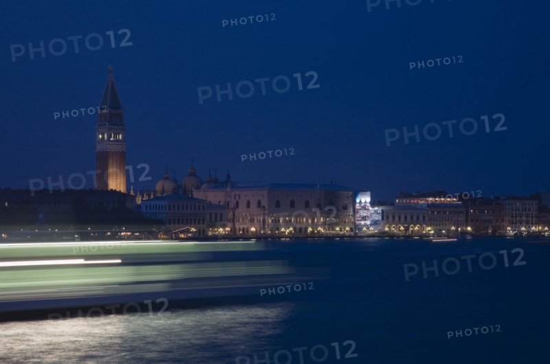 Venice, Veneto, Italy. Giudecca island Early evening view towards illuminated Campanile of St. Mark with vaporetto water bus to Murano and Burano in blurred motion in foreground with light trails reflected in water of the canal. Italy Italia Italian Venice Veneto Venezia Europe European City Giudecca Island St saint Mark Marks Campanile Vaporetto Water Bus Blur Blurred Movement Streak Streaks Streaked Light Lights Transport Night Illuminated Destination Destinations Nite Religion Southern Europe Warm Light