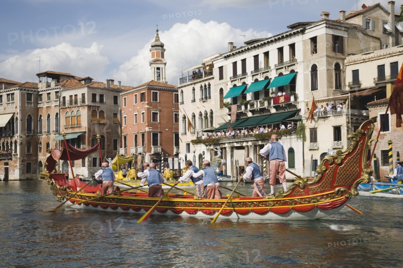 Venice, Veneto, Italy. Participants in the Regata Storico historical Regatta held each September rowing red and gold decorated gondola and wearing traditional costume approaching the Rialto bridge with onlookers gathered on the balconies of canalside buildings behind. Teams represent Sestiere districts of Venice in traditional races. Italy Italia Italian Venice Veneto Venezia Europe European City Regata Regatta Gondola Gondola Gondolas Gondolier Boat Architecture Exterior Water Blue Classic Classical Clouds Cloud Sky Destination Destinations History Historic Holidaymakers Older Southern Europe Tourism Tourist
