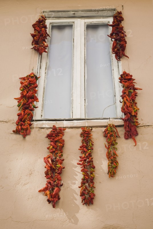Kusadasi, Aydin Province, Turkey. Strings of red and orange chili peppers hung up to dry in summer sunshine from window frame of house in the old town. Turkey Turkish Eurasia Eurasian Europe Asia Turkiye Aydin Province Kusadasi Chilli Chilli Chillis Chillis Chilie Chillies Pepper Peppers Capsicum Capsicums Window Drying Red Color Destination Destinations European Middle East South Eastern Europe Western Asia