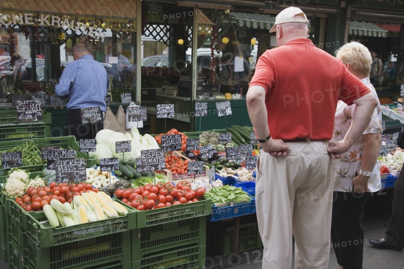 Vienna, Austria. The Naschmarkt. Tourists looking at display on fresh produce stall in front of glass fronted shop front including aubergines tomatoes sweetcorn and cauliflower. Austrian Destination Destinations European Holidaymakers Osterreich Store Tourism Viena Western Europe Wien