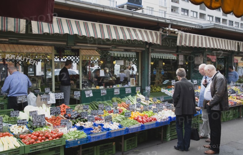 Vienna, Austria. The Naschmarkt. Customers at fresh produce stall looking at display that includes tomatoes aubergines leeks courgettes and mushrooms. Austria Austrian Republic Vienna Viennese Wien Europe European City Capital Naschmarkt Market Display Fresh Food Fruit Vegetables Veg Vegetable Stall Shop Store Destination Destinations Osterreich Viena Western Europe