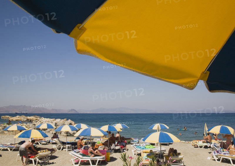Kos, Dodecanese Islands, Greece. People sunbathing on beach outside Kos Town with sun loungers blue and yellow striped parasols and people in shallow water of rock enclosed bay beyond. Part framed by underside of parasol in foreground. Greece Greek Europe European Vacation Holiday Holidays Travel Destination Tourism Ellas Hellenic Docecanese Kos Beach Sun Sunbathers Sunbathing Loungers Tourists Parasols Sea Umbrellas Blue Yellow Color Destination Destinations Ellada Holidaymakers Sand Sandy Beaches Tourism Seaside Shore Tourist Tourists Vacation Southern Europe