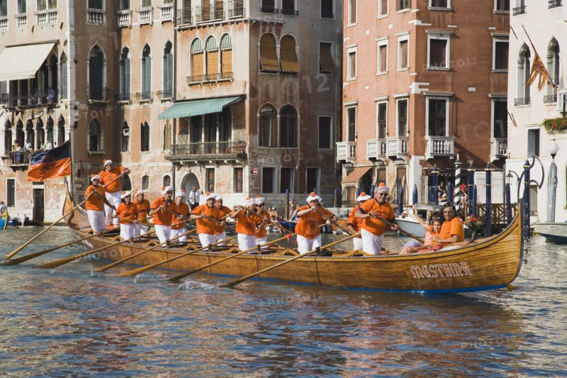 Venice, Veneto, Italy. Regatta Storico historical annual regatta. Team of rowers wearing orange and white costume passing canalside buildings with onlookers watching from balconies. Italy Italia Italian Venice Veneto Venezia Europe European City Regata Regatta Gondola Gondola Gondolas Gondolier Boat Architecture Exterior Water Destination Destinations History Historic Holidaymakers Southern Europe Tourism Tourist