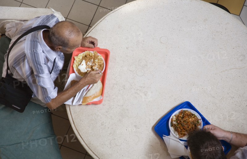 Budapest, Pest County, Hungary. Looking down on two men having lunchtime meal seated at circular table in Nagy Vasarcsarnok the Central Market. Hungary Hungarian Europe European East Eastern Buda Pest Budapest City Nagy Vasarcsarnok Central Market Food Eating lunch Lunchtime Meal Men Table Seated Aerial Trays 2 Eastern Europe Male Man Guy