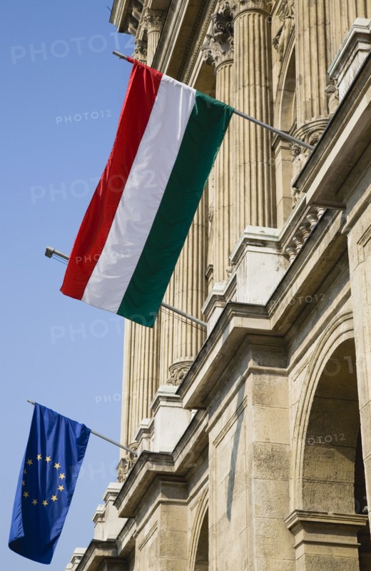 Budapest, Pest County, Hungary. Hungarian and EU flags fying from facade of building on the River Danube in Pest. Hungary Hungarian Europe European East Eastern Buda Pest Budapest City Architecture Facade Exterior Flag Flags EEC EU Union Tricolor Tricolour Blue Destination Destinations Eastern Europe European Union