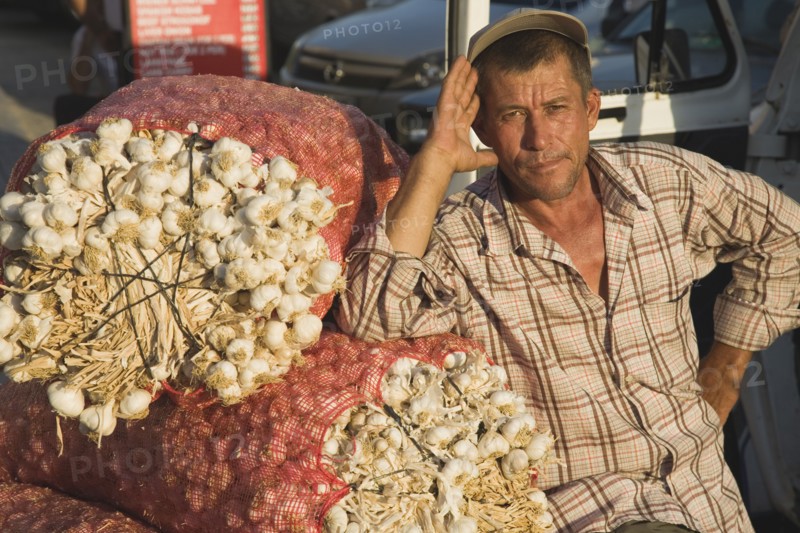 Kusadasi, Aydin Province, Turkey. Stallholder at weekly market leaning on sacks of garlic in the late afternoon summer sunshine. Turkey Turkish Eurasia Eurasian Europe Asia Turkiye Aydin Province Kusadasi Market markets Stall Vendor Man Male Garlic Sacks Destination Destinations European Male Men Guy Middle East One individual Solo Lone Solitary South Eastern Europe Western Asia