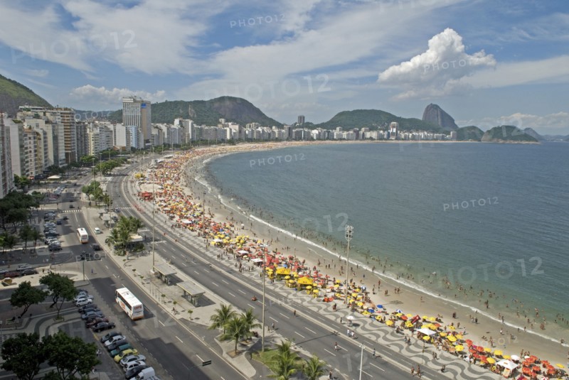 Rio de Janeiro, Brazil. Copacabana. The full sweep of Avenue Atlantica mosiac sidewalk with crowds on the beach and in the sea. Umbrellas hotels tour buses Sugarloaf Mountain blue sea and sky with puffy cloud. American Brasil Brazilian Destination Destinations Latin America Latino Sand Sandy Beaches Tourism Seaside Shore Tourist Tourists Vacation South America Southern Sunbather Travel Water