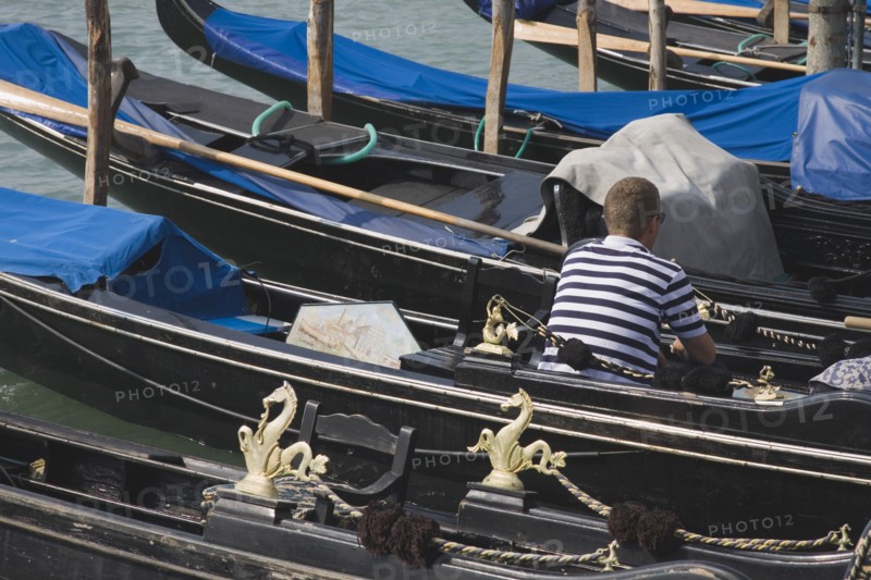 Venice, Veneto, Italy. Gondolier wearing striped shirt using mobile phone sitting in one of a line of moored gondolas with back to camera. Italy Italia Italian Venice Veneto Venezia Europe European City Water Shirt Striped Stripes Stripe Stripe Cell Mobile Phone Gondolier Gondolas Gondola 1 Cellular One individual Solo Lone Solitary Single unitary Southern Europe