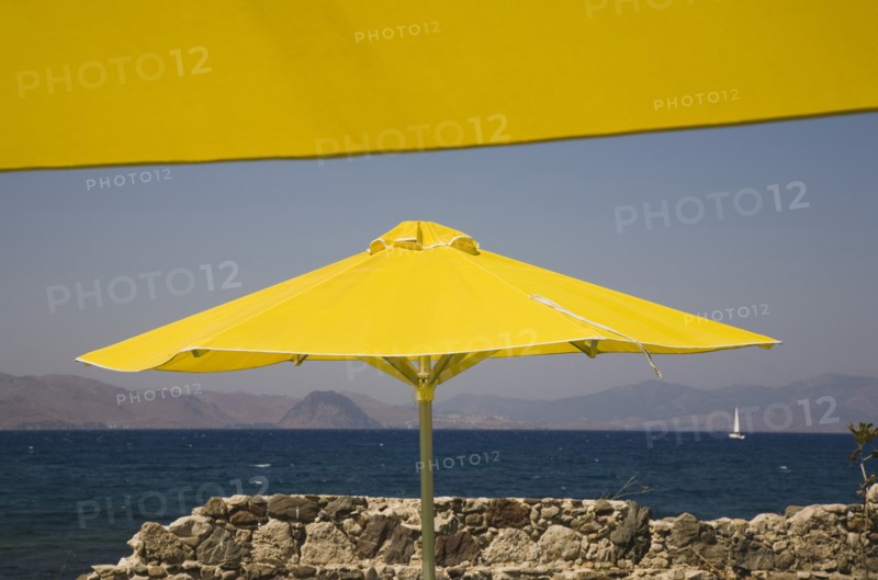 Kos, Dodecanese Islands, Greece. Bright yellow parasols on beach outside Kos Town with crumbling stone wall in foreground and view towards distant coastline beyond. Greece Greek Europe European Vacation Holiday Holidays Travel Destination Tourism Ellas Hellenic Docecanese Kos Sea Umbrella Umbrellas Parasol Parasols Yellow Blue Color Destination Destinations Ellada Sand Sandy Beaches Tourism Seaside Shore Tourist Tourists Vacation Southern Europe Water