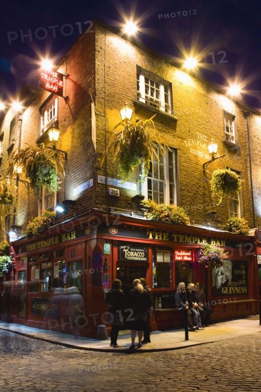 Ireland, County Dublin, Dublin City, Temple Bar Pub illuminated at night with people walking past on the cobbled streets of the area on the south bank of the Liffey River.