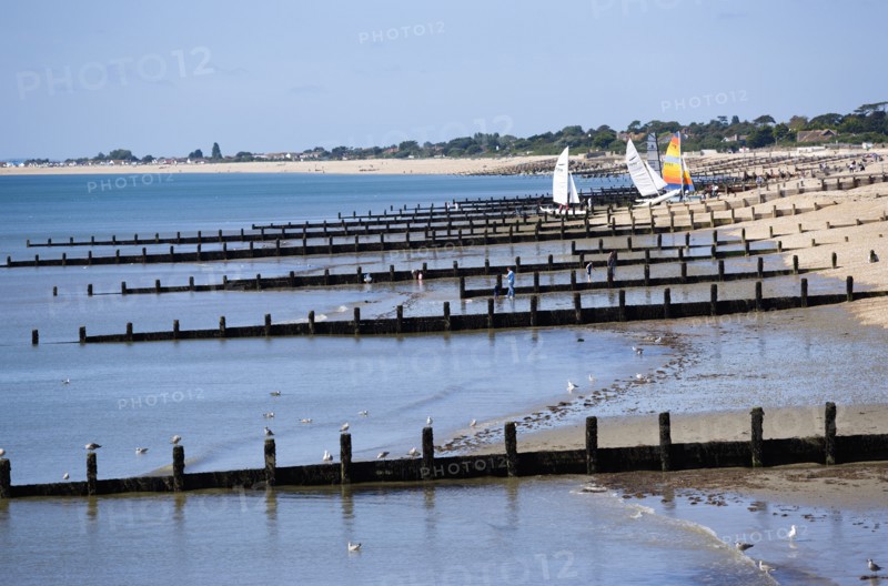 England, West Sussex, Bognor Regis, Wooden groynes at low tide used as sea defences against erosion of the shingle pebble beach with people on the sand by the waterline and small sailing boats on the beach.
