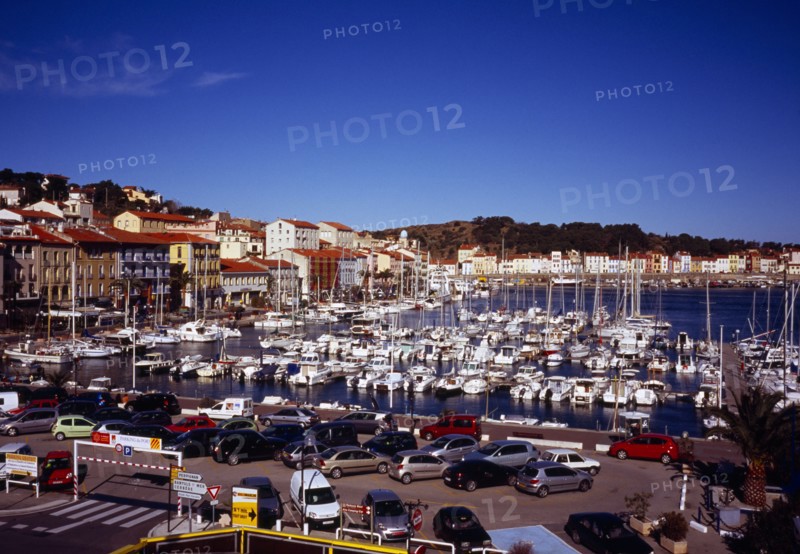 France, Languedoc-Roussillon, Pyrenees Orientales, Port-Vendres.  Inner harbour with moored boats  quay and quayside buildings with car park in foreground. Hillside part visible beyond.