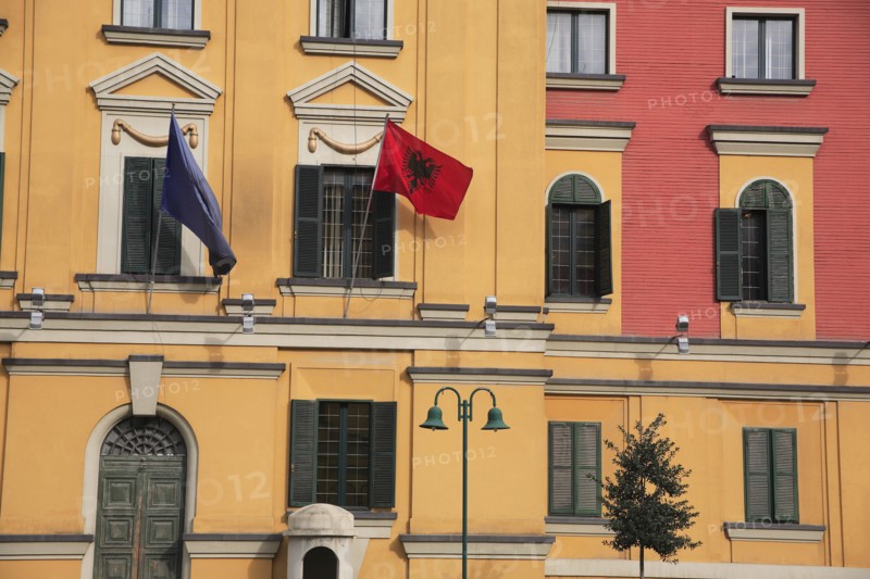 Albania, Tirane, Tirana, Part view of pink and yellow exterior facade of government buildings in Skanderbeg Square flying flags including double headed eagle emblem. Multiple windows with green painted shutters and green painted  arched doorway.