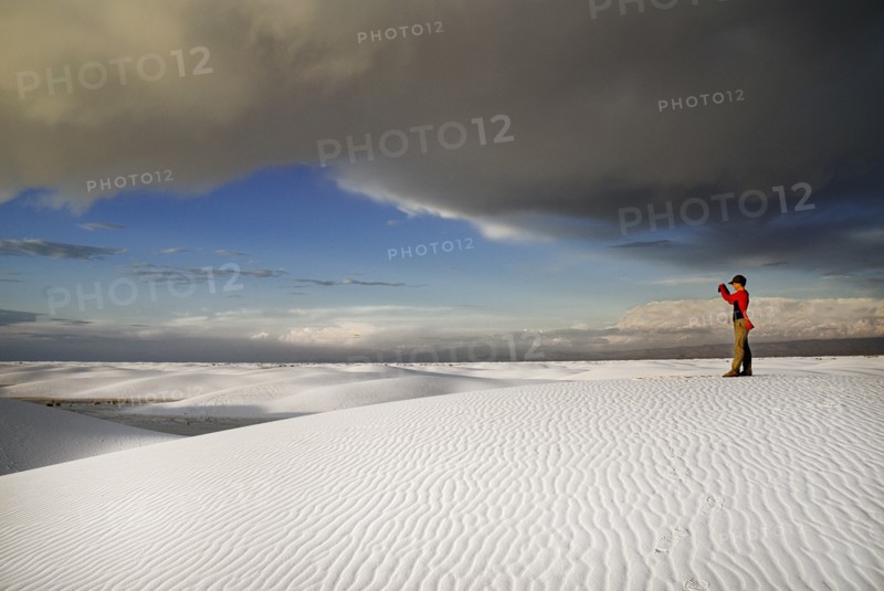 USA, New Mexico, Otero County, White Sands National Monunment. Landscape of white sand with dramatic cloudscape above. Single figure in mid-ground taking photograph. 
Photo : Hugh Rooney