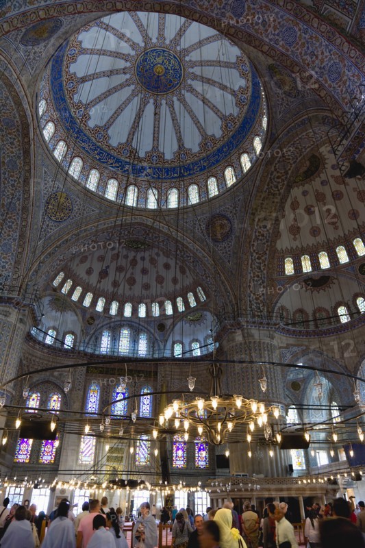 Turkey, Istanbul, Sultanahmet Camii The Blue Mosque interior with sightseeing tourists by a chandelier below the decorated domes with stained glass windows beyond. 
Photo : Paul Seheult