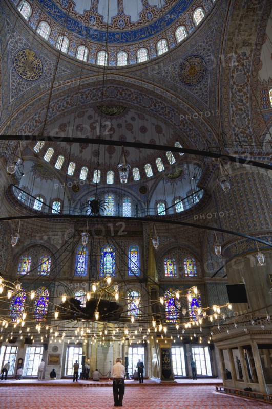Turkey, Istanbul, Sultanahmet Camii The Blue Mosque interior with people at prayer beneath chandeliers and decorated domes above and stained glass windows beyond. 
Photo : Paul Seheult