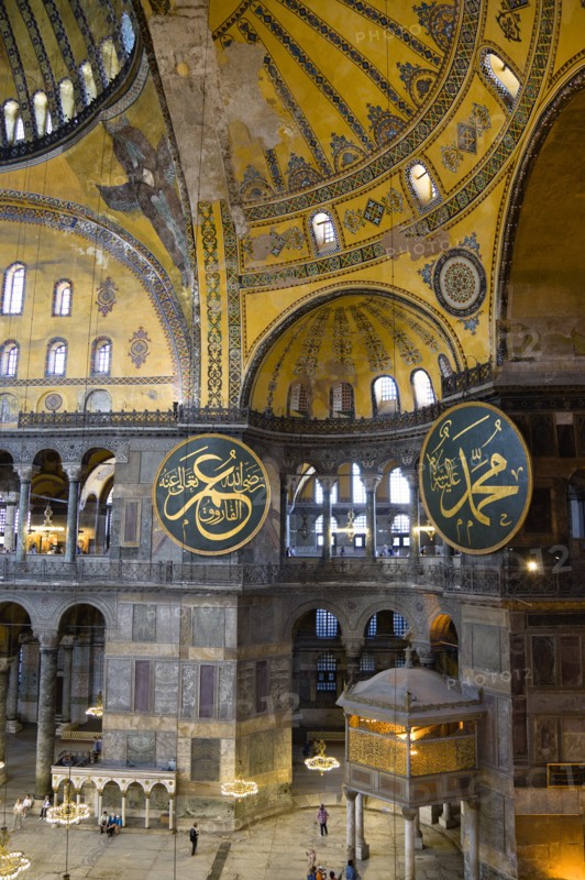 Turkey, Istanbul, Sultanahmet Haghia Sophia Sightseeing tourists beneath the dome with murals and chandeliers in the Nave of the Cathedral with calligraphic roundels of Arabic Koran texts above the Lodge of The Sultan. 
Photo : Paul Seheult