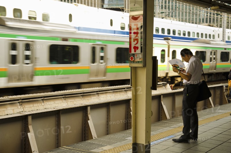 Japan, Honshu, Tokyo, Commuter on platform cooling himself with paper fan whilst waiting for his train. 
Photo : Jon Burbank
